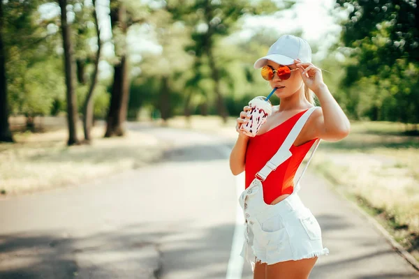 Retrato de sexy chica de moda elegante con gafas de sol redondeadas, traje de baño rojo, gorra de béisbol blanca, pantalones cortos con batido al aire libre. —  Fotos de Stock