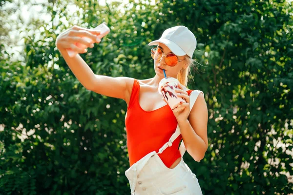 Hezká mladá fena s šálkem studeného nápoje s úsměvem a přičemž selfie při stání na rozmazaném pozadí keřů v parku — Stock fotografie