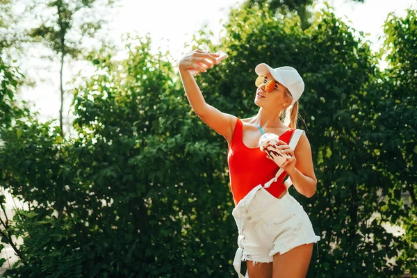 Bastante joven hembra con taza de bebida fría sonriendo y tomando selfie mientras está de pie sobre un fondo borroso de arbustos en el parque —  Fotos de Stock
