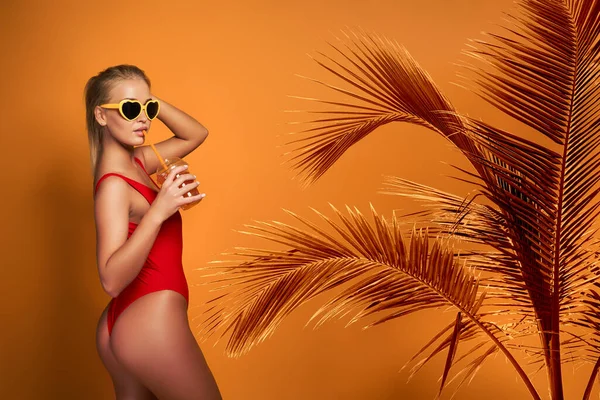 Schöne junge Frau in Badebekleidung und Sonnenbrille, die ein Glas frisches Getränk hält, während sie auf gelbem, orangefarbenem Hintergrund steht — Stockfoto