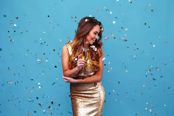 Ung elegant kvinna i gyllene glitter klänning. kväll makeup innehav glas champagne och firar över blå studio bakgrund med konfetti. — Stockfoto