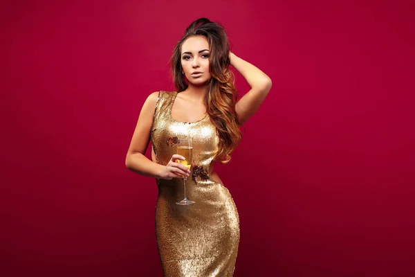 Giovane donna elegante in abito glitter dorato. trucco serale in possesso di un bicchiere di champagne e festeggiare su sfondo rosso studio. — Foto Stock