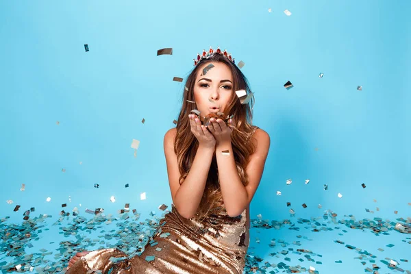 Retrato de la joven hermosa chica en vestido de oro y la corona posando sobre fondo azul sopla confeti de sus manos a la cámara — Foto de Stock