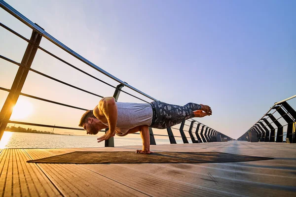 Ajuste al hombre haciendo una parada de una mano mientras practica yoga solo cerca del océano contra el cielo al atardecer o al amanecer —  Fotos de Stock