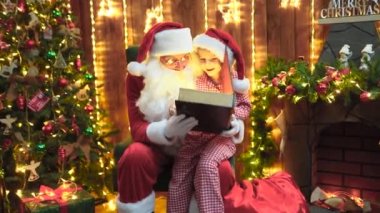 Küçük kız Noel Baba 'nın dizlerinin üstüne çöküp hediye kutusunu ya da sihirli kitabı açıyor.