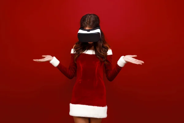 Удивительная юная Санта-девушка в рождественском платье, смотрящая в наушники указывая руки в сторону изолированы на красном фоне стены. — стоковое фото