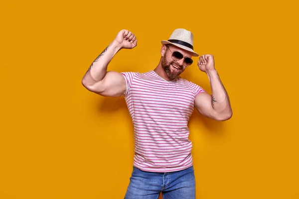 Imagen del hombre tatuado barbudo excitado en gafas de sol y sombrero haciendo gesto ganador fondo amarillo aislado. — Foto de Stock