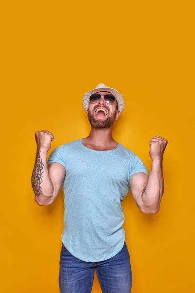 Imagen del hombre tatuado barbudo excitado en gafas de sol y sombrero haciendo gesto ganador fondo amarillo aislado. — Foto de Stock