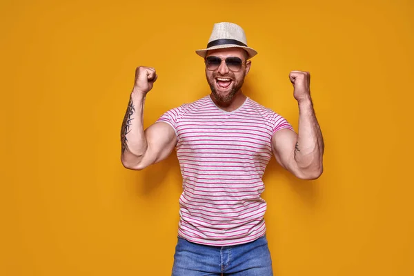 Imagen del hombre tatuado barbudo excitado en gafas de sol y sombrero haciendo gesto ganador fondo amarillo aislado. — Foto de Stock