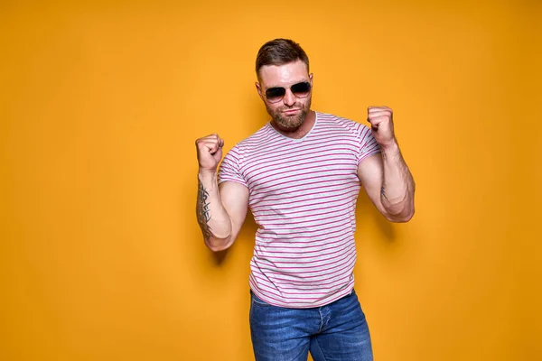 Imagen de hombre barbudo excitado en gafas de sol haciendo gesto ganador aislado fondo amarillo — Foto de Stock