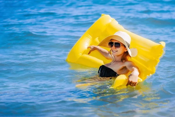 Letni obraz kobiety pływającej na żółtym dmuchanym materacu w oceanie. Noszący słomkowy kapelusz, okulary przeciwsłoneczne. — Zdjęcie stockowe