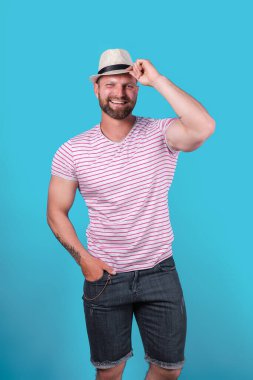 Modaya uygun, kaslı, sakallı, saman şapkalı gülümseyen adam stüdyoda mavi arka planda poz veriyor.