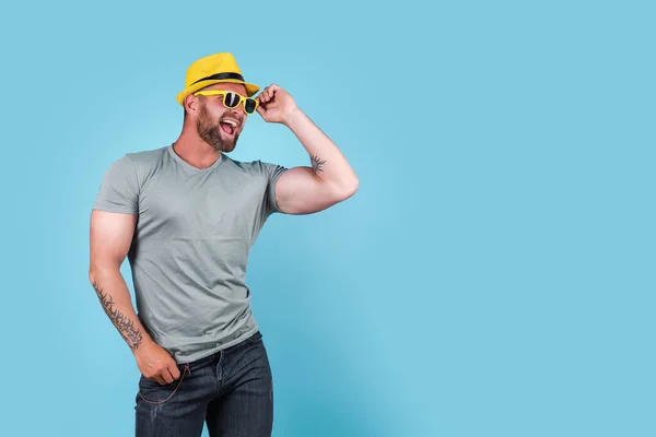 Modischer muskulöser bärtiger emotionaler Mann mit gelbem Strohhut posiert im Studio vor blauem Hintergrund — Stockfoto