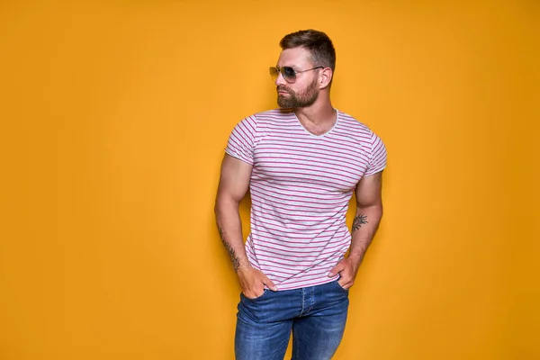 Gespierde zelfverzekerde man met baard in shirt, jeans en stijlvolle zonnebril staand in studio over gele achtergrond — Stockfoto