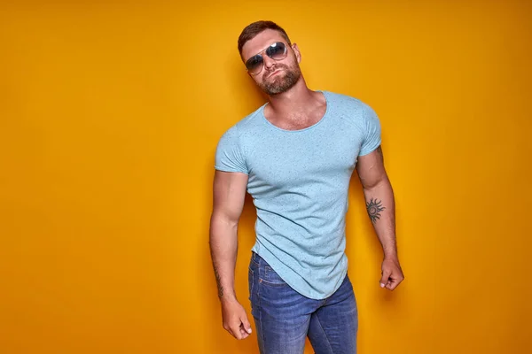 Musculoso hombre barbudo con confianza en camisa, jeans y gafas de sol con estilo de pie en el estudio sobre fondo amarillo — Foto de Stock