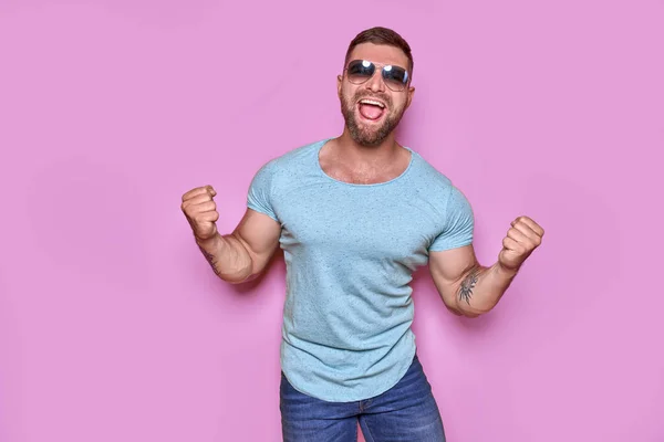 Imagen del hombre tatuado barbudo excitado en gafas de sol gesto ganador aislado fondo rosa. — Foto de Stock