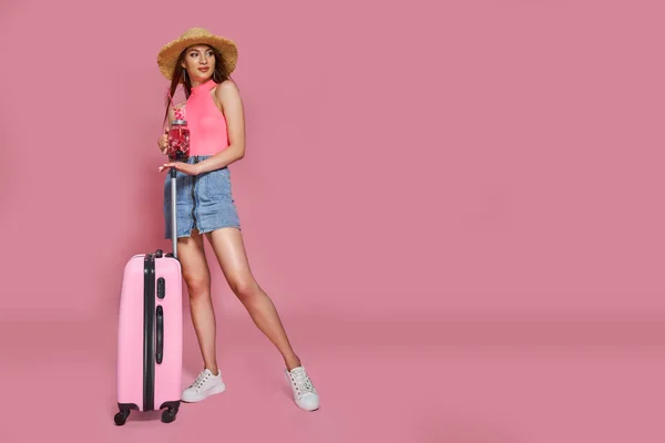Туристична жінка в літньому повсякденному одязі, що тримає свіжі напої та валізу на рожевому фоні — стокове фото