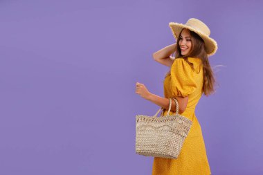 Sarı elbiseli, yaz hasır şapkalı, pastel menekşe arka planda izole edilmiş çantalı güzel gülümseyen genç kadın.. 