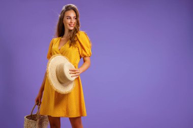 Sarı elbiseli, yaz hasır şapkalı, pastel menekşe arka planda izole edilmiş çantalı güzel gülümseyen genç kadın.. 