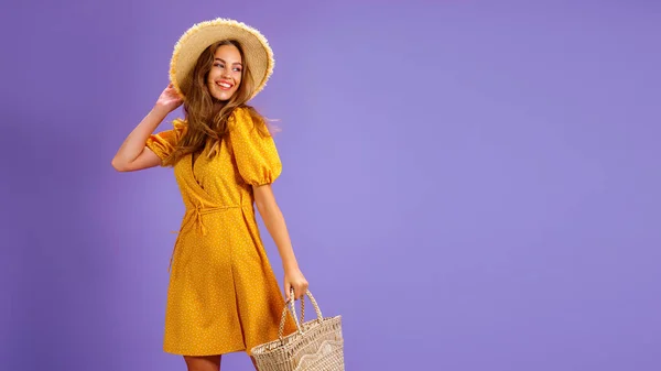 Mooie glimlachende jonge vrouw in gele jurk, zomer strohoed, tas geïsoleerd op pastel violette achtergrond. — Stockfoto