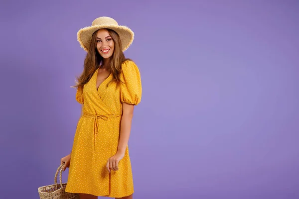 Hermosa joven sonriente en vestido amarillo, sombrero de paja de verano, bolsa aislada sobre fondo violeta pastel. —  Fotos de Stock