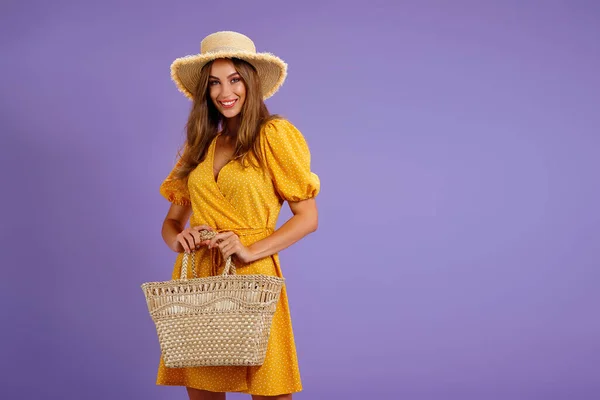 Красива усміхнена молода жінка в жовтій сукні, літній солом'яний капелюх, сумка ізольована на пастельному фіолетовому фоні . — стокове фото