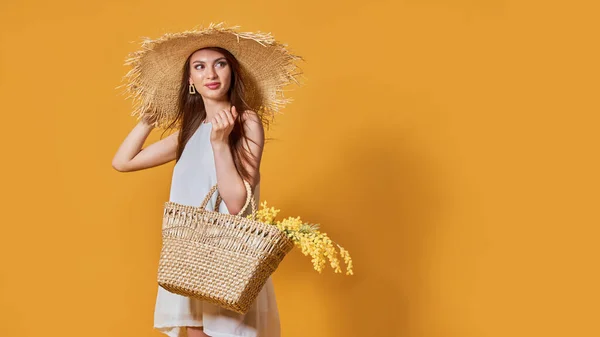 Щаслива жінка в літній білій сукні, солом'яний капелюх з купою квітів у сумці позує на жовтому студійному фоні — стокове фото