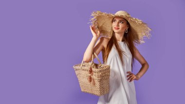 Beyaz elbiseli, yaz hasır şapkalı, güneş gözlüklü, pastel menekşe arka planda izole edilmiş saman çantalı güzel bir kadın.. 