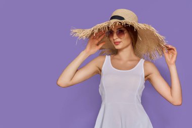 20 'li yaşlardaki genç moda kadınının menekşe arka planda saman şapka ve güneş gözlüğü taktığı fotoğraf.