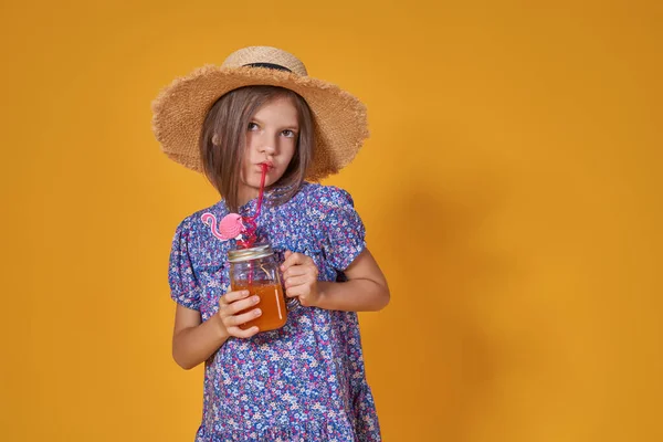 Little girl in a straw hat and sunglasses is smiling on a yellow background. child girl in a blue dress is holding a drink glass on a yellow background — Stock Photo, Image