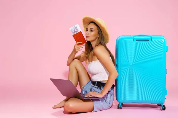 Een jonge vrouw met een hoed, spijkerbroek en wit topje heeft een paspoort met documenten in haar hand en kijkt naar laptop terwijl ze naast een blauwe koffer zit. — Stockfoto