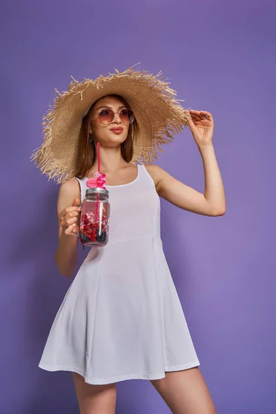 Mooi meisje in stro hoed en zonnebril met een verfrissende zomer cocktail op paarse achtergrond. — Stockfoto