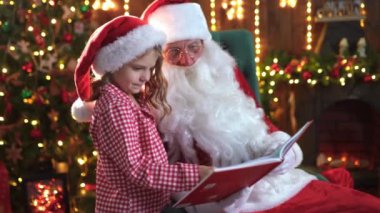 Pijamalı küçük kız ve Noel Baba sihirli kitap okuyor.