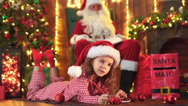 Rolig flicka i pyjamas och Santa hatt skriver brev till Santa nära julgran. — Stockvideo