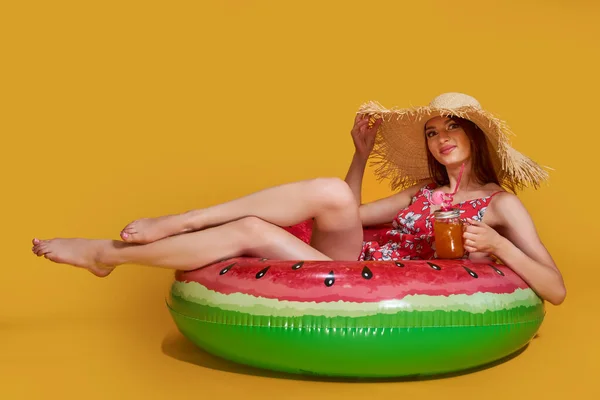 Beautiful girl in summer dress, straw hat sit on air mattress in circle shape of watermelon with cocktail, yellow studio background. — Stock Photo, Image