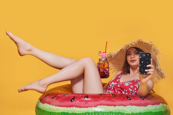 Beautiful girl in summer dress, straw hat with inflatable ring shape of watermelon with fresh cocktail, talking online by mobile phone, yellow studio background. — Stock Photo, Image