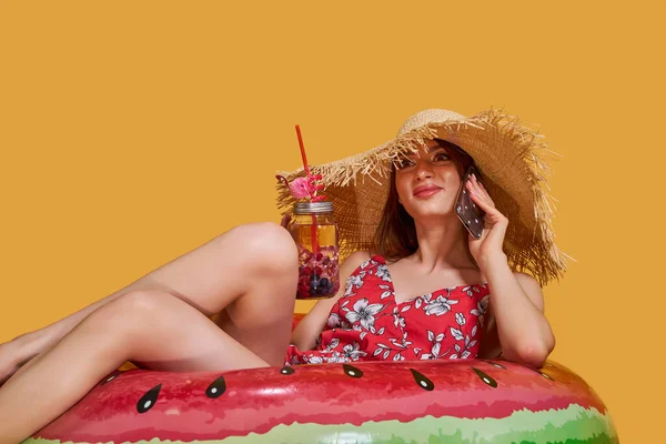 Mooi meisje in de zomer jurk, stro hoed zitten op luchtbed in cirkelvorm van watermeloen en drink cocktail, praten op mobiele telefoon, gele studio achtergrond. — Stockfoto