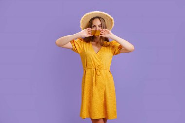 Sarı elbiseli ve şapkalı neşeli beyaz kadın elinde mor arka planda izole edilmiş banka kartı tutuyor.