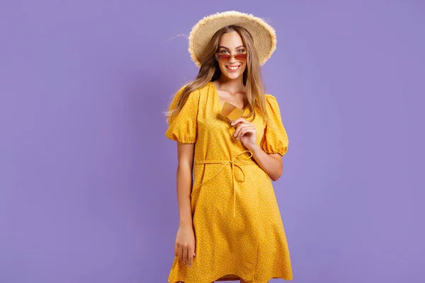 身穿黄色衣服头戴草帽的快乐的白人年轻女子手持用紫色背景隔开的银行卡 — 图库照片