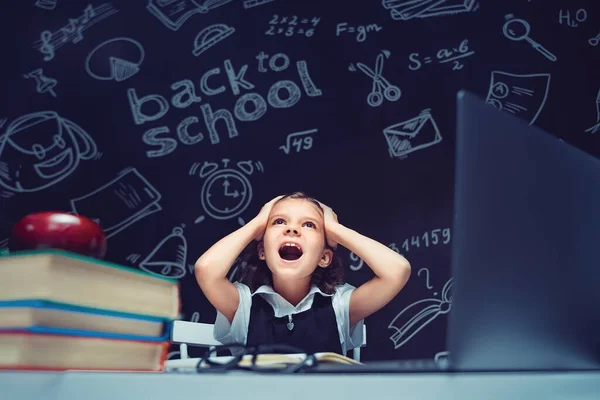 Schoolgirl κάθεται στο τραπέζι με φορητό υπολογιστή, βιβλία και ένα chalkboard με τύπους σχολείο στο παρασκήνιο. Εργασίες σπίτι έκπληκτος να βάλει τα χέρια της στο κεφάλι, ανοίγοντας το στόμα ευρύ — Φωτογραφία Αρχείου