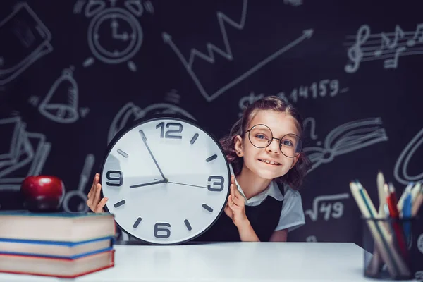 Leerling meisje met grote klok op het zwarte schoolbord in de klas. — Stockfoto