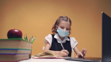 Kız tıp maskeli, okul üniformalı bir kız, bir masada oturuyor ve bir dizüstü bilgisayarla internetten okuyor.. 