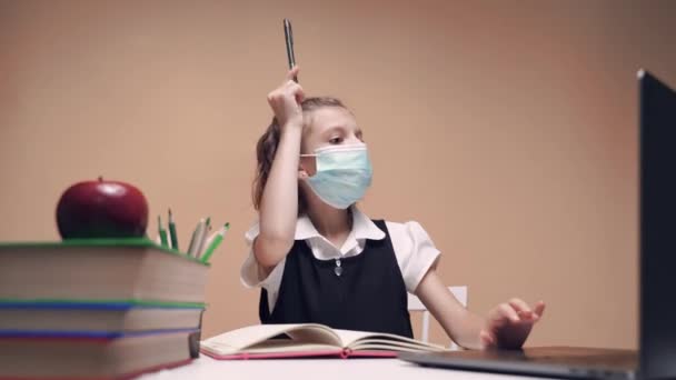 Chica es una colegiala con una máscara médica, uniforme escolar, se sienta en un escritorio y estudia en línea a través de un ordenador portátil, va a escribir, pero deja caer un bolígrafo. — Vídeos de Stock