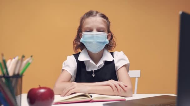 Kız tıp maskeli, okul üniformalı bir kız, bir masada oturuyor ve bilgisayarla internetten çalışıyor, kamera hareket ediyor.. — Stok video