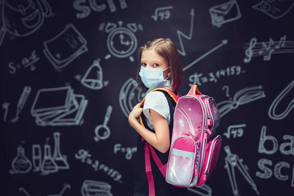 Liten flicka i skolan uniform bär medicinsk mask med ryggsäck på svarta tavlan bakgrund — Stockfoto