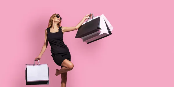 Mujer joven en un vestido negro y gafas de sol sostiene bolsas de compras sobre un fondo rosa —  Fotos de Stock