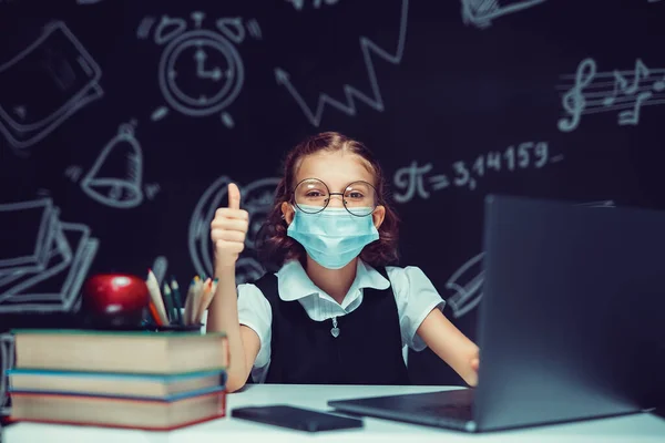 Junior student bär medicinsk mask online virtuell inlärning språk klass på bärbar dator visar tummen upp. — Stockfoto