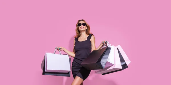 Fiatal nő fekete ruhában és napszemüveg tartja bevásárló táskák rózsaszín háttér — Stock Fotó