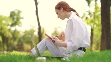 Çimenlerde oturan bir kız notlarla bir şeyler yazıyor ve dizüstü bilgisayarla çalışıyor.