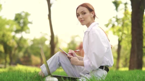 Egy lány ül a fűben, jegyzetel, laptoppal dolgozik és kamerába néz. — Stock videók
